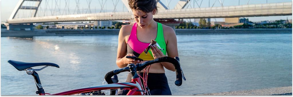 junge Radfahrerin, die einen Energieriegel isst und sich am Flussufer erholt, nachdem sie mit dem Fahrrad gefahren ist. Gesunder Lebensstil und Sportkonzept.