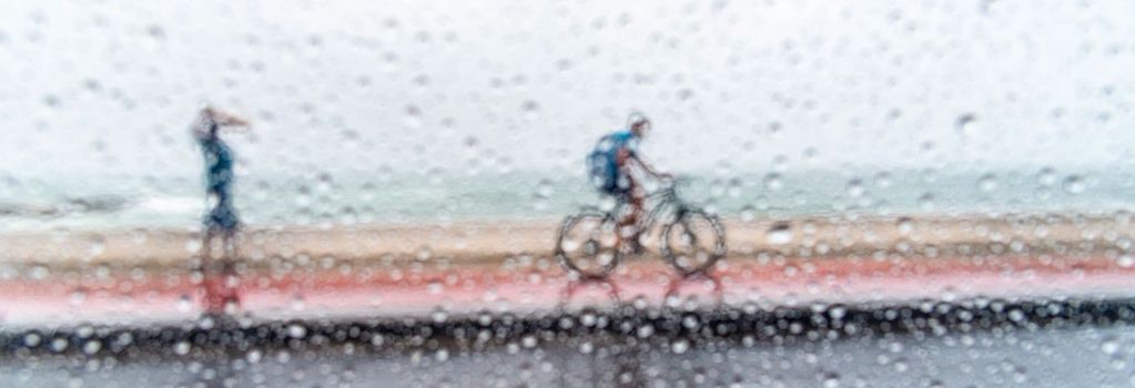 Der Blick fällt durch eine Glasscheibe, die voller Regentropfen ist: Dahinter sehen wir einen Radfahrer, im Regen auf einem Radweg fährt.