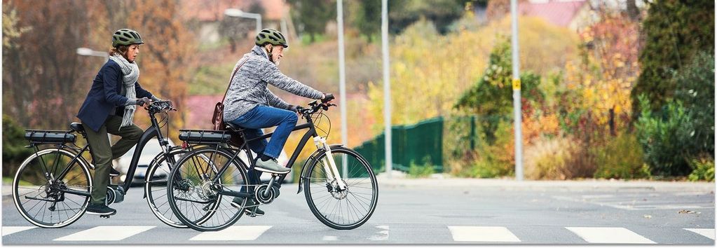 Seniorenpaar fährt auf Elektrofahrrädern über eine Straße. Im Hintergrund ist eine herbstliche Szenerie zu sehen.