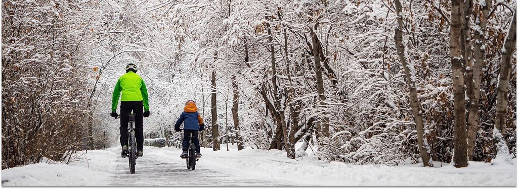 Ein Vater fährt mit seinem Sohn auf Fahrrädern auf einem schneebedeckten Weg durch einen Winterwald. Sie tragen Helme und ihr Fahrrad sowie ihre Kleidung sind wintertauglich.