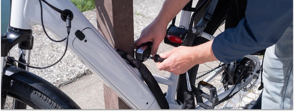 Detailaufnahme: Jemand schließt sein E-Bike mit einem Kettenschloss an einen Pfeiler an.