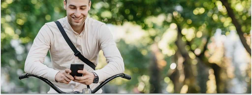 Ein junger Mann sitzt auf seinem Fahrrad und schaut auf ein Smartphone in seinen Händen, die Arme auf dem Lenker abgelegt.