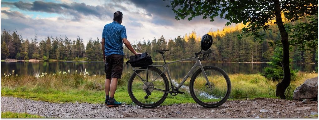 Cube Nuroad C:62 EX steht in ländlicher Umgebung an einem See. Daneben steht ein Mann, der sich am E-Bike abstützt.
